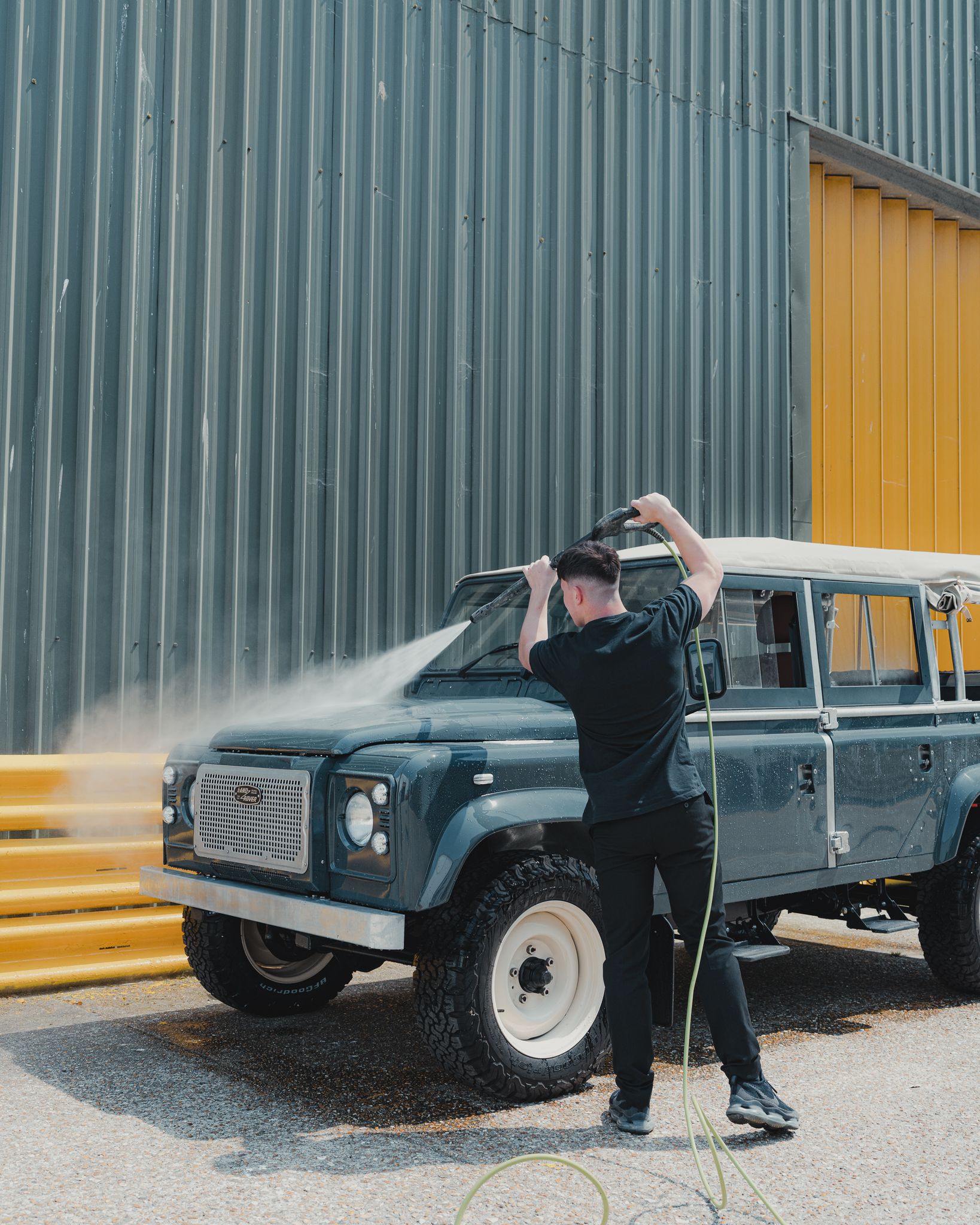 A man uses a high-pressure hose to wash a large SUV beside a corrugated building wall.